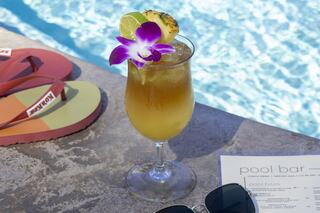 A tropical cocktail garnished with a flower and pineapple sits by a pool, alongside sunglasses and colorful flip-flops.