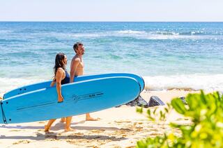 A couple walks along the beach carrying blue surfboards, with a bright ocean and sunny sky in the background, enjoying a perfect day.