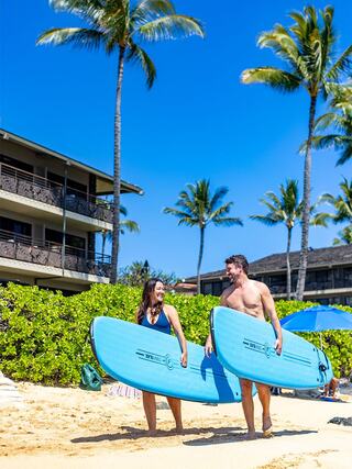 A man and woman, carrying surfboards, walk along a sunny beach surrounded by palm trees and beachfront accommodations. Happy vibes abound!