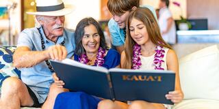 A smiling family enjoys looking at a photo book together, wearing floral leis in a bright, inviting indoor setting.