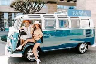 Two women in stylish outfits pose beside a vintage blue van labeled "PASÉA," with palm trees and modern architecture in the background.