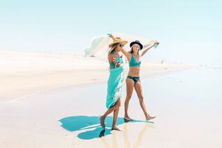Two smiling women walk along a sunlit beach, one draped in a turquoise towel, wearing hats and swimwear, enjoying a carefree day by the ocean.