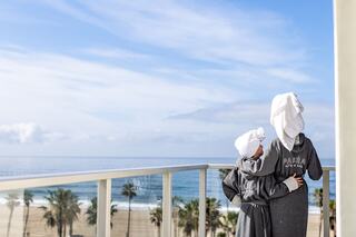 A person and a child in matching robes and towel headdresses stand on a balcony, gazing at the serene ocean and beach view.