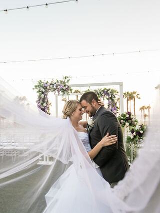 A joyful couple embraces under a floral arch at sunset, with the bride's veil flowing gently in the breeze, exuding romance and happiness.