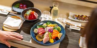 A beautifully arranged platter of sushi and sashimi garnished with vibrant vegetables, accompanied by drinks and elegant dishware.