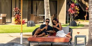 A couple enjoys a cozy moment outdoors, sharing food and drinks on a lounge bed, surrounded by lush greenery and a relaxed atmosphere.