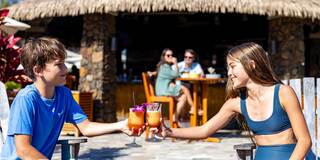 Two children are cheerfully toasting with colorful drinks in a sunny outdoor setting, surrounded by tropical elements and relaxed atmosphere.