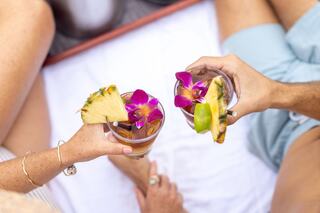 Two hands hold tropical drinks adorned with flowers and fruit, set against a backdrop of relaxed legs on a blanket.