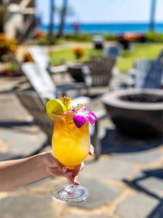 A hand holds a vibrant tropical cocktail adorned with a lime wedge and orchid, set against a backdrop of sunlit loungers and ocean views.