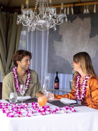 A couple sits at a elegantly set table adorned with flower petals, enjoying each other's company under a chandelier. A bottle of wine is visible.