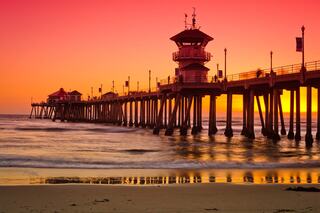 A vibrant sunset paints the sky in hues of orange and pink, illuminating a pier with tall pillars and a charming structure at the end.