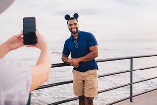 A smiling man in a blue polo and shorts poses with Mickey Mouse ears while someone takes his photo by the ocean.