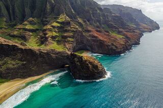Lush green cliffs rise dramatically above turquoise waters, framing a secluded sandy beach along a rugged coastline.