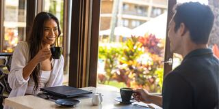 A woman enjoys coffee while smiling at a man in a sunny cafe, surrounded by lush greenery. A relaxed and intimate atmosphere prevails.