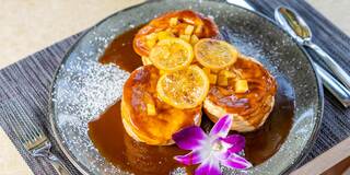 Three golden pastries topped with caramelized fruit slices, drizzled with sauce, and garnished with a bright purple flower.