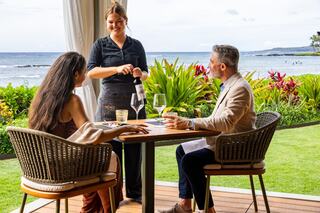 A couple enjoys a meal by the ocean while a waitress serves wine, surrounded by vibrant greenery and a serene coastal view.