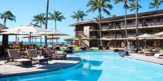 A sunny poolside scene with loungers and umbrellas, surrounded by palm trees and a multistory building, near a beautiful ocean view.