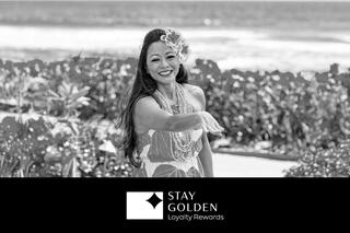 A joyful woman in a colorful outfit smiles and gestures warmly, surrounded by lush greenery and ocean waves.