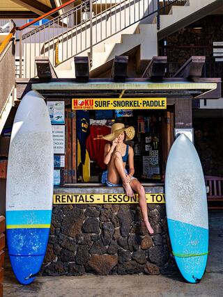 A woman in a bikini and sunhat relaxes at a surf rental shop, with surfboards on either side against a stone wall backdrop.