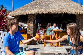 Two children toast with tropical drinks in a sunny outdoor setting, surrounded by greenery and a vibrant social atmosphere.