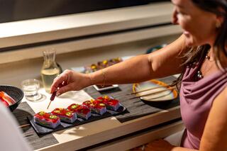 A woman delicately drizzles sauce over beautifully arranged sushi on a sleek dining table, enjoying a moment of culinary artistry.