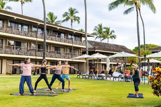 A group of five people practice yoga outdoors on a green lawn, surrounded by palm trees and a resort-like setting.