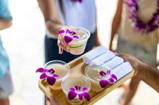 A wooden tray holds drinks garnished with purple orchids, alongside rolled towels, as hands reach to share refreshments in a tropical setting.