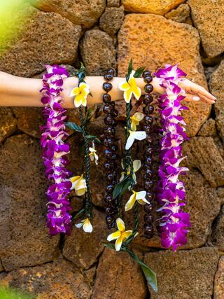 A hand showcases vibrant floral leis in purple and white against a textured stone wall, highlighting tropical beauty and cultural adornment.