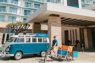 A man stands next to a vintage blue van and a tricycle outside a modern hotel entrance, featuring stylish architecture and greenery.