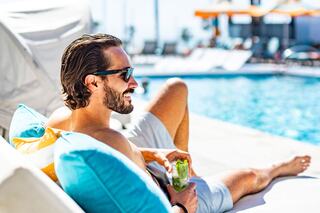A man relaxes by the pool on a sunbed, wearing sunglasses and enjoying a drink, surrounded by colorful cushions and bright sunshine.