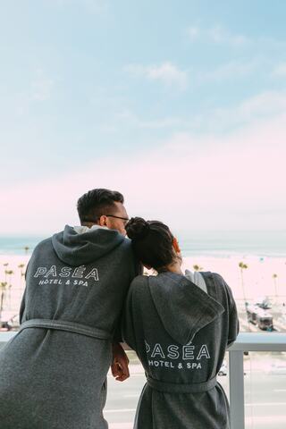 A couple in matching robes gazes at a beach view from a balcony, enjoying a serene moment together. Palm trees frame the scene.