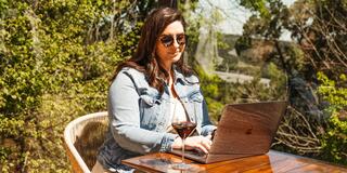 A woman in sunglasses works on a laptop outdoors, enjoying a cocktail at a table surrounded by lush greenery.