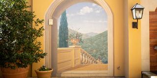 A painted archway offers a scenic view of hills and greenery, framed by a warm yellow wall and adorned with potted plants and a lantern.