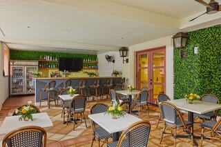 A quaint café space features tables set with floral arrangements, a bar lined with beverages, and lush green walls for a welcoming ambiance.