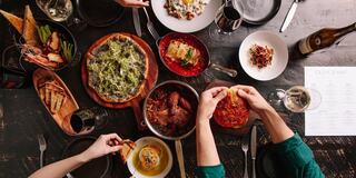 A vibrant table filled with various delicious dishes, wine, and drinks, showcasing a feast with hands reaching for food.