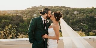 A couple embraces and kisses on a scenic rooftop, surrounded by lush greenery and a serene landscape at sunset.