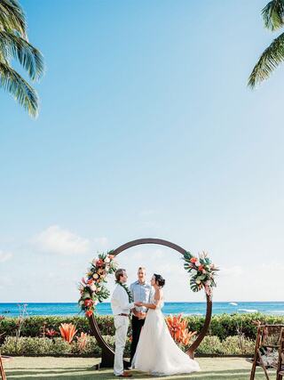 A couple exchanges vows on a sunny beach with a floral arch and ocean backdrop, surrounded by tropical greenery and blue skies.