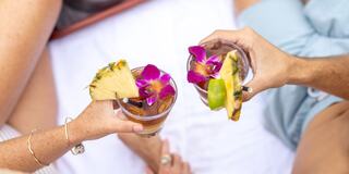 Two hands hold tropical cocktails adorned with flowers and fruit, set against a soft white backdrop, suggesting a relaxed, vacation vibe.