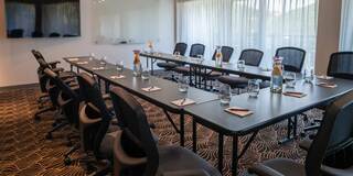 A modern meeting room features a long table with chairs, water bottles, and notepads, illuminated by natural light through large windows.