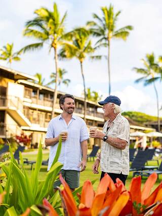 Two men enjoy drinks while walking through a tropical setting, surrounded by vibrant flowers and palm trees, with a relaxed resort atmosphere.