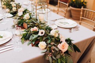 A beautifully set dining table adorned with greenery and soft floral arrangements, featuring elegant dinnerware and glassware.