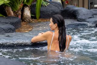 A woman with long hair relaxes in a hot spring, leaning against rocks, surrounded by lush greenery. Tranquility in nature.