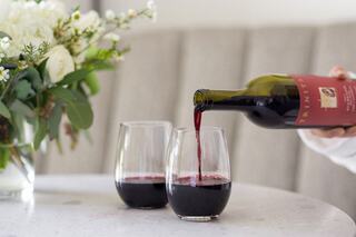 A bottle of Trinitas Cellars red wine is being poured into two glasses on a table, accompanied by a bouquet of white flowers in a vase.