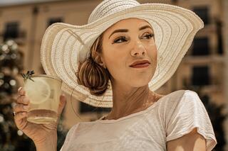 A woman in a wide-brimmed hat holds a refreshing drink with lemon, gazing thoughtfully in a sunny outdoor setting.