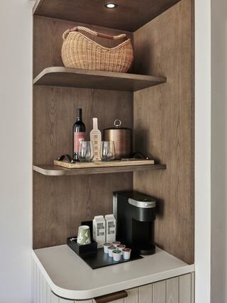 A modern kitchenette features wooden shelves with a basket, wine, glasses, a pot, and a coffee maker with assorted coffee pods.