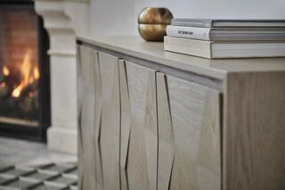 A textured wooden cabinet with geometric patterns sits beside a cozy fireplace, topped with books and a decorative gold bowl.