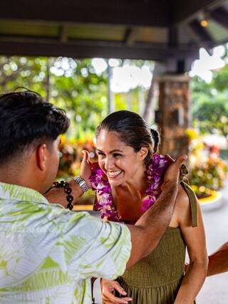 A joyful woman receives a flower lei from a smiling man, set against a lush, tropical backdrop with vibrant greenery.