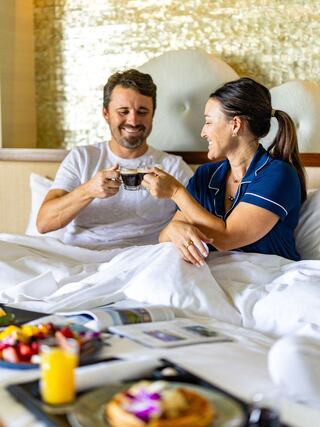 A couple enjoys coffee in bed, smiling and sharing a joyful moment, with a delicious breakfast spread beside them.