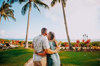 A couple stands embraced, admiring a tropical landscape with palm trees and colorful flowers, overlooking a serene ocean.