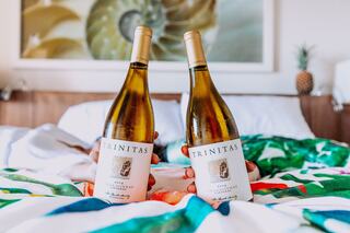 Two bottles of Trinitas Chardonnay are held above a colorful bedspread, set against a backdrop of a cozy room with decor accents.